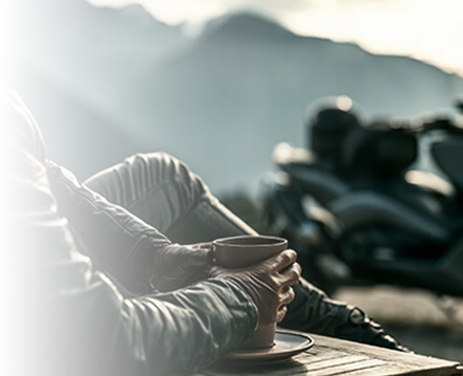 A rider holding a coffee at a mountain stop, with the 318 parked behind