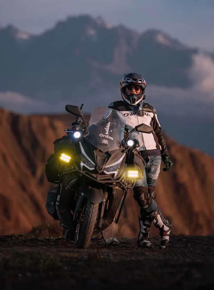 A rider in full gear beside a 318 at dusk, lights on
